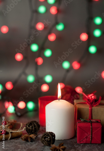 Red and white festive candles burn with a yellow flame on a table among gifts in craft paper  rattan balls  cinnamon sticks and anise stars on the background of a multi-colored Christmas garland