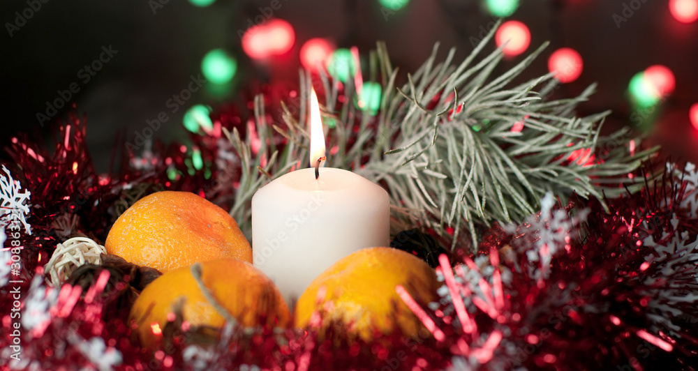A white candle burns with flame on a table among gifts in craft paper, rattan balls, spruce wreath, tangerines, cinnamon sticks on a background of multi-colored Christmas garland