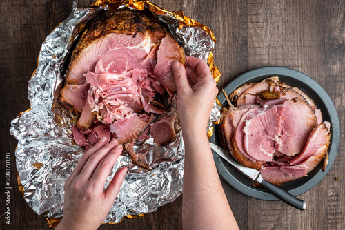 Woman’s hand pulling pieces of ham off a Spiral cut glazed and cooked ham in a foil wrapper on a wood table, knife and black plate with slices photo