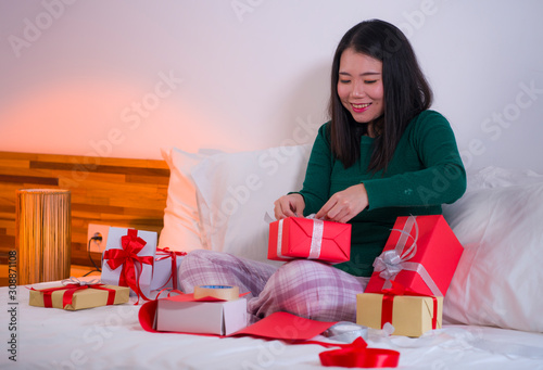 Christmas holiday lifestyle portrait of young happy and sweet Asian American woman sitting on bed preparing xmas presents wrapping gift boxes and adding ribbons