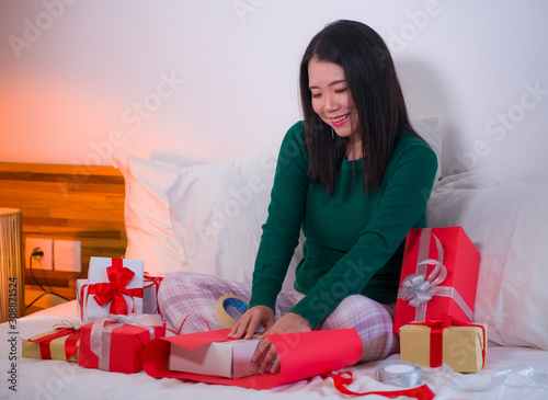 Christmas holiday lifestyle portrait of young happy and sweet Asian Japanese woman sitting on bed preparing xmas presents wrapping gift boxes and adding ribbons