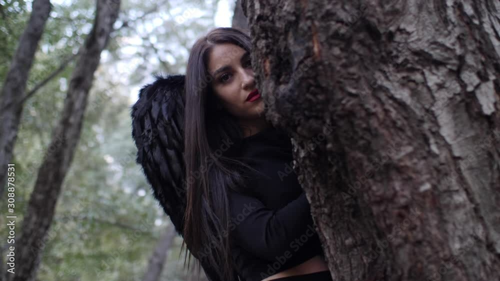 Beautiful gothic model with black angel wings peaks out from behind a tree in the forest. Beautiful gothic model with black angel wings peaks out from behind a tree in the forest.