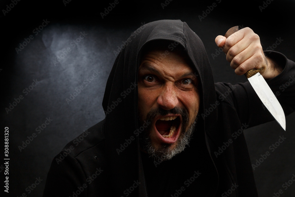 Portrait of a bald man with a beard in a black hood with a knife on a dirty gray background. Maniac. Portrait of a bald man with a beard in a black hood with a knife on a dirty gray background. Maniac.