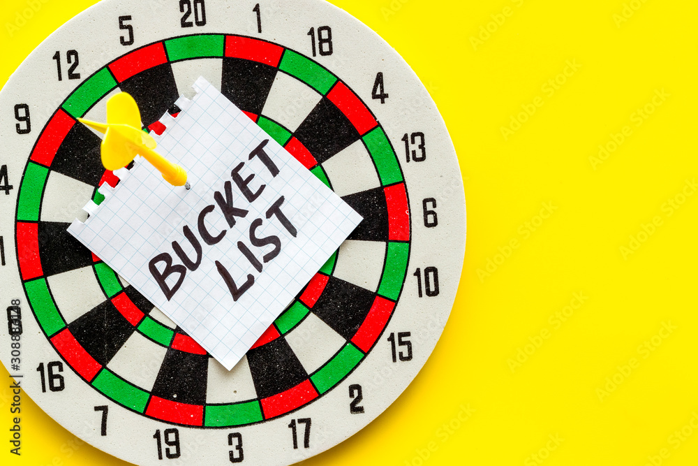 Bucket list attached by dart to dart board on yellow background top-down copy space Bucket list attached by dart to dart board on yellow background top-down copy space