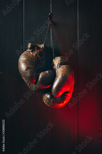 Brown retro leather boxing gloves hanging on a black wooden wall in a red light. © a_khachatryan