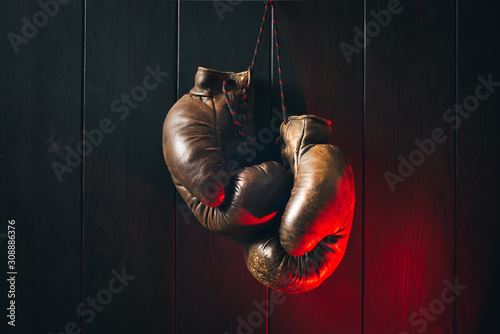 Old brown boxing gloves, hanging on black wooden wall in dramatic red lighting. Copy space. © a_khachatryan