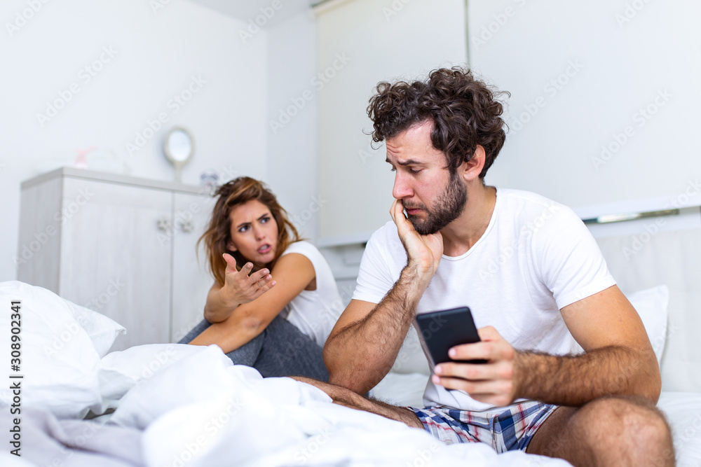 Young couple is sitting on the bed. The guy is looking at something on his smart phone. The girl is offended at him. She is looking at him annoyed and frustrated by boyfriend on the phone Young couple is sitting on the bed. The guy is looking at something on his smart phone. The girl is offended at him. She is looking at him annoyed and frustrated by boyfriend on the phone