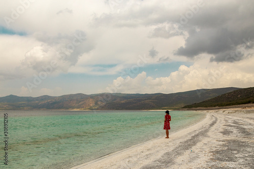 woman model walking through Salda lake, Turkey with her red dress and blue hat. Turqouise water and beach with cloudy sky