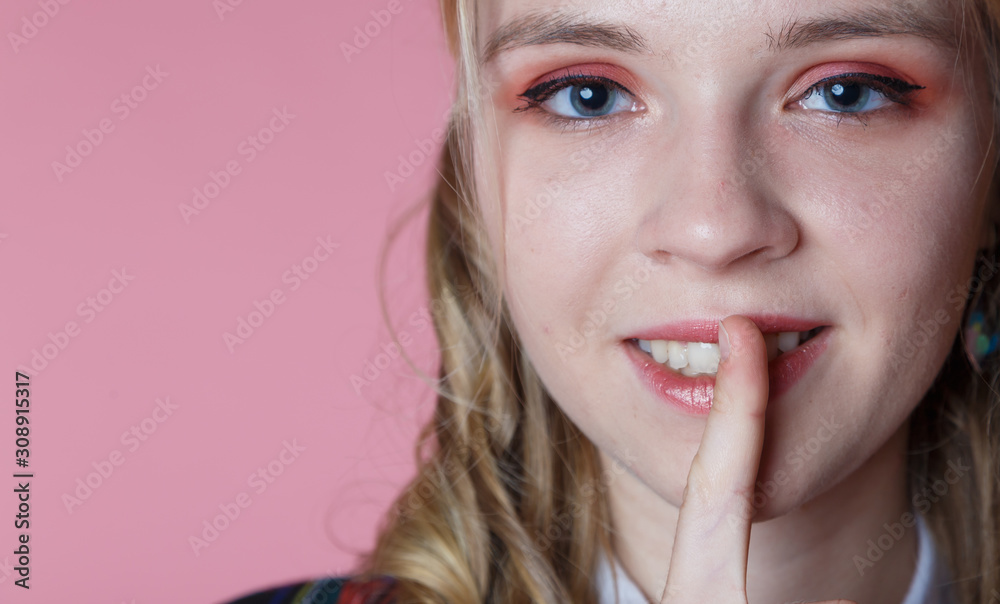 young blonde curly haired girl holds her finger to her lips on a pink background close-up, concept of secret and silence. selective focus young blonde curly haired girl holds her finger to her lips on a pink background close-up, concept of secret and silence. selective focus