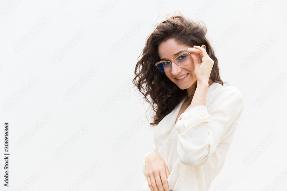 Positive female professional adjusting glasses and smiling at camera. Wavy haired young woman in casual shirt standing isolated over white background. Corporate female portrait concept
