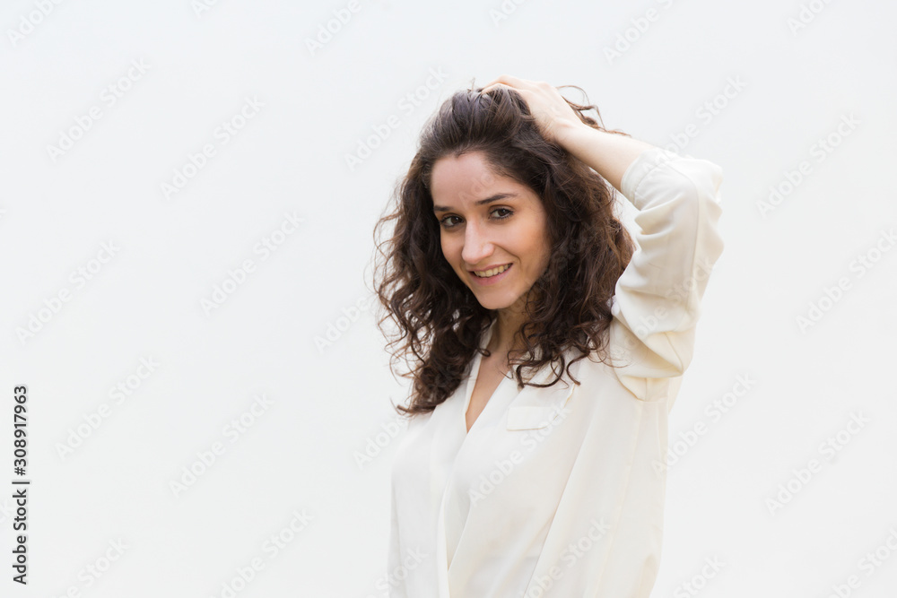Happy positive beautiful woman touching hair, looking at camera, smiling. Wavy haired young woman in casual shirt standing isolated over white background. Hair care concept Happy positive beautiful woman touching hair, looking at camera, smiling. Wavy haired young woman in casual shirt standing isolated over white background. Hair care concept