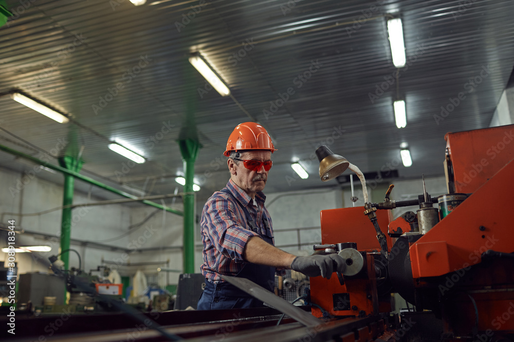 Mature manual worker wearing protective eyeglasses standing at lathe holding the lever and controlling the machine Mature manual worker wearing protective eyeglasses standing at lathe holding the lever and controlling the machine