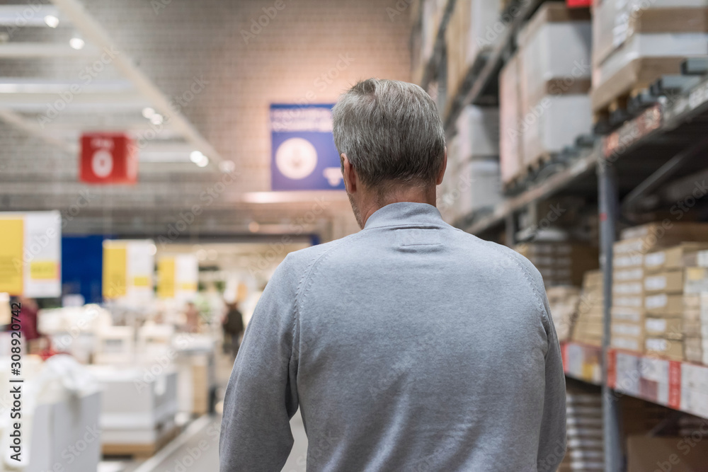 Tall gray-haired man shopper walking with trolley in a large hardware store before christmas. Back view Tall gray-haired man shopper walking with trolley in a large hardware store before christmas. Back view