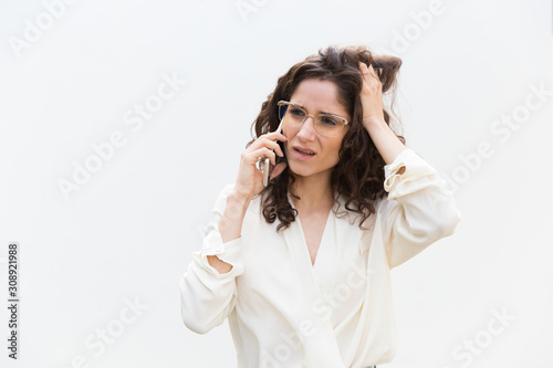 Stressed woman in glasses speaking on cell phone, touching head in confusion. Wavy haired young woman in casual shirt standing isolated over white background. Bad mobile connection concept
