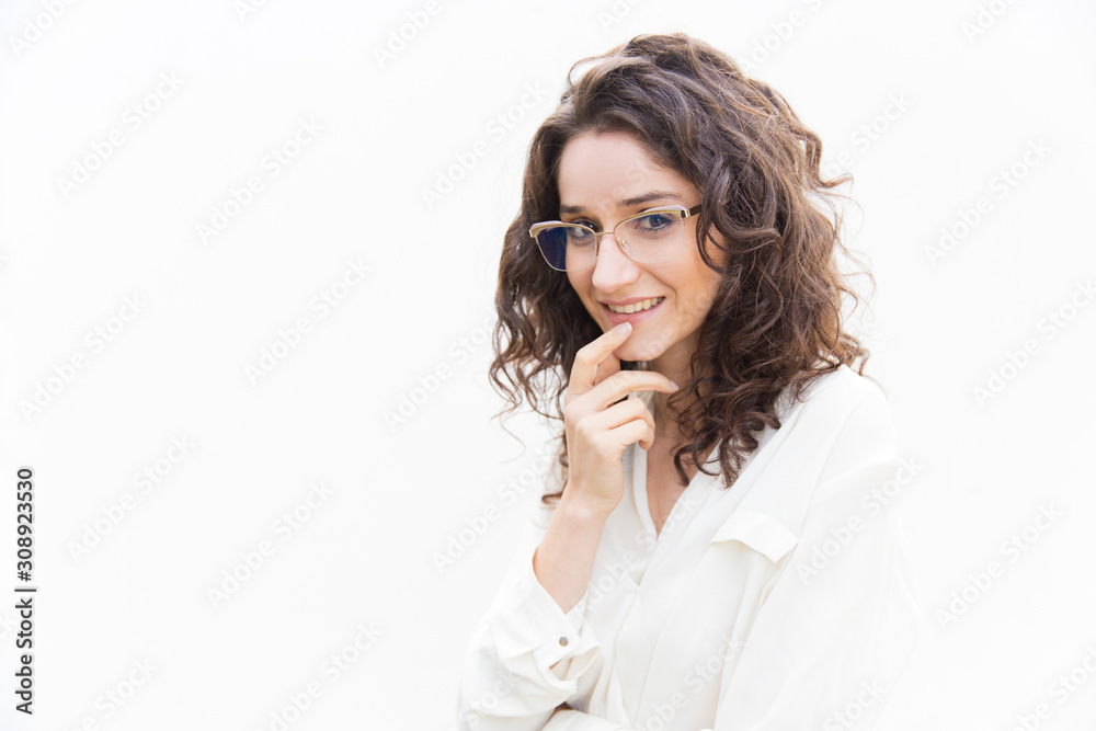 Pensive female customer in glasses smiling, touching chin, looking at camera. Wavy haired young woman in casual shirt standing isolated over white background. Thinking concept