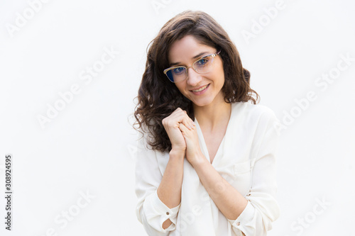 Positive female professional with clasped hands smiling at camera. Wavy haired young woman in casual shirt standing isolated over white background. Female portrait concept