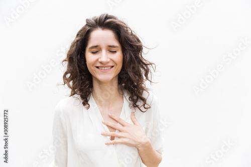 Happy grateful pretty woman with closed eyes applying hand to chest. Wavy haired young woman in casual shirt standing isolated over white background. Relief or gratitude concept