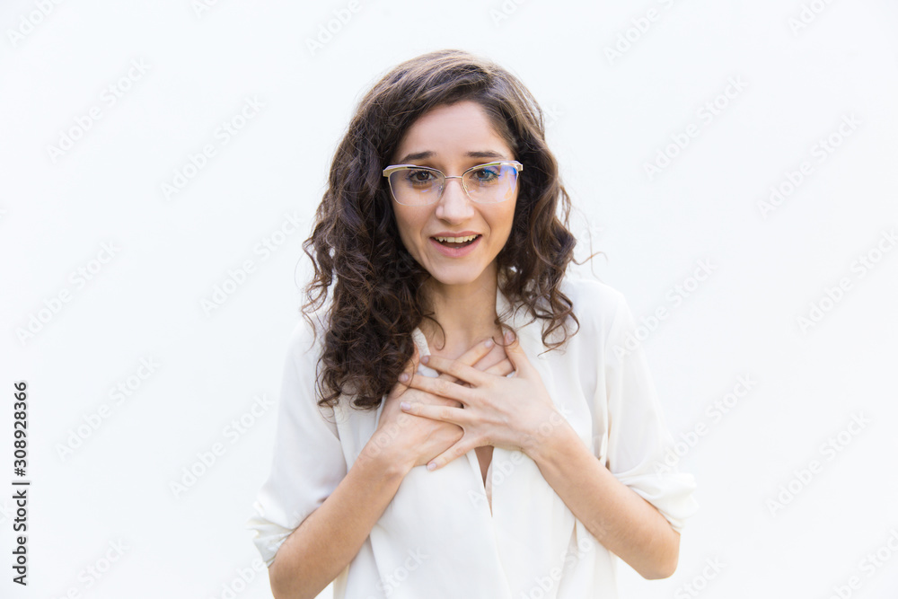 Happy amazed woman in glasses gasping and applying hands to chest. Wavy haired young woman in casual shirt standing isolated over white background. Surprise concept