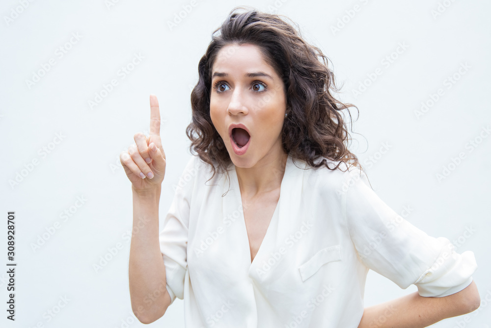 Extremely amazed woman gasping, pointing finger up, looking away. Wavy haired young woman in casual shirt standing isolated over white background. Aha moment concept Extremely amazed woman gasping, pointing finger up, looking away. Wavy haired young woman in casual shirt standing isolated over white background. Aha moment concept