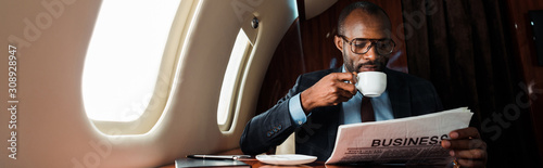 Panoramic shot of African American businessman in glasses reading business newspaper while holding cup in private plane photo