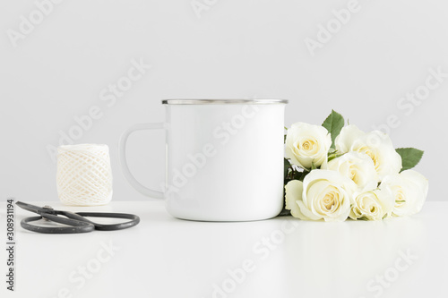 Enamel mug mockup with a bouquet of roses and workspace accessories on a white table.