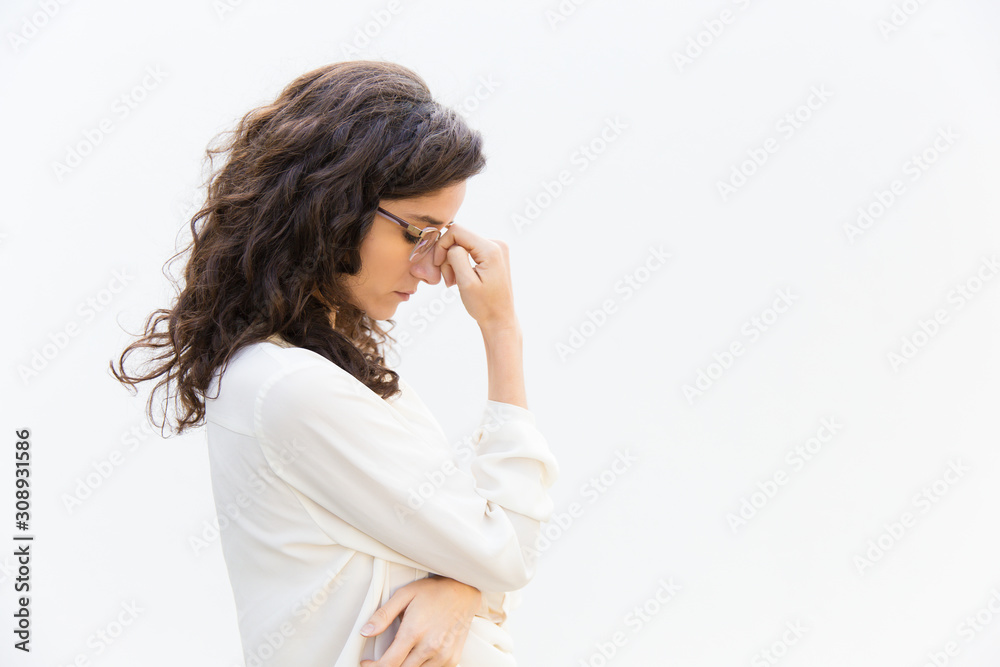 Side of tired sad office worker in glasses with closed eyes leaning head on hand. Wavy haired young woman in casual shirt standing isolated over white background. Business problem concept Side of tired sad office worker in glasses with closed eyes leaning head on hand. Wavy haired young woman in casual shirt standing isolated over white background. Business problem concept