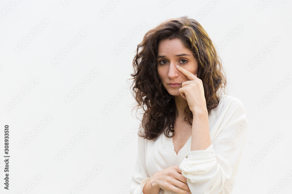 Sad unhappy woman removing tears from face, looking at camera. Wavy haired young woman in casual shirt standing isolated over white background. Crying concept Sad unhappy woman removing tears from face, looking at camera. Wavy haired young woman in casual shirt standing isolated over white background. Crying concept