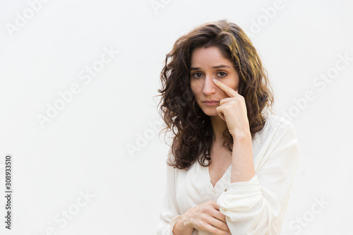Sad unhappy woman removing tears from face, looking at camera. Wavy haired young woman in casual shirt standing isolated over white background. Crying concept