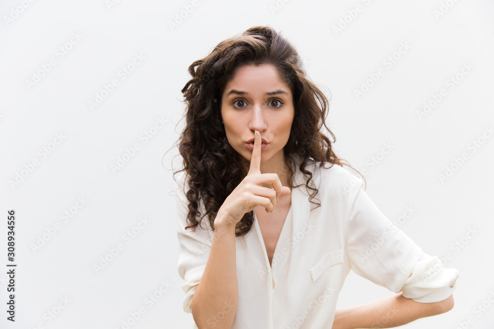 Serious concerned woman showing shh gesture. Wavy haired young woman in casual shirt standing isolated over white background. Keeping silence concept Serious concerned woman showing shh gesture. Wavy haired young woman in casual shirt standing isolated over white background. Keeping silence concept