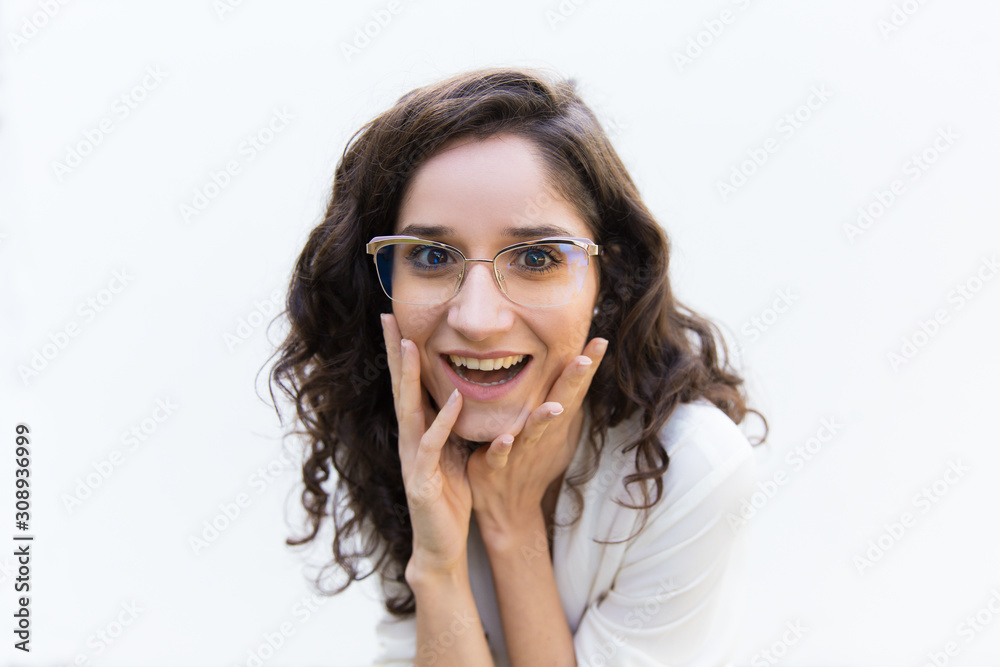 Excited amazed female professional in glasses touching face and staring at camera. Wavy haired young woman in casual shirt standing isolated over white background. Sharing news concept