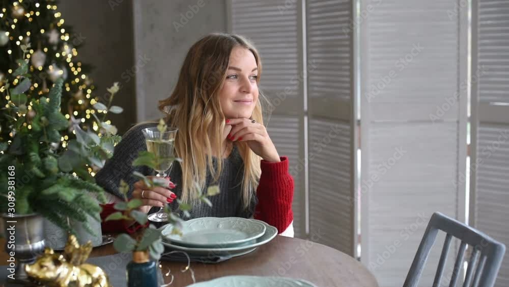 Happy beautiful blonde with long hair is sitting at the table, looking out the window and drinking champagne in the living room decorated for Christmas Happy beautiful blonde with long hair is sitting at the table, looking out the window and drinking champagne in the living room decorated for Christmas