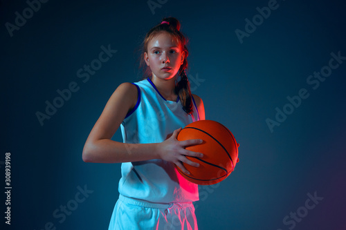 Strength.Young caucasian female basketball player on blue studio background in neon light, motion and action. Concept of sport, movement, energy and dynamic, healthy lifestyle. Posing confident. © master1305