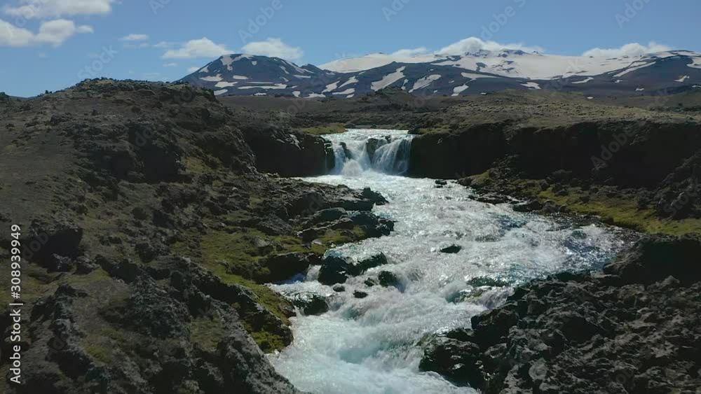 Flying a drone up river with pure and cold water towards a waterfall in the highlands of Iceland. Snow capped mountains in the background. Flying a drone up river with pure and cold water towards a waterfall in the highlands of Iceland. Snow capped mountains in the background.