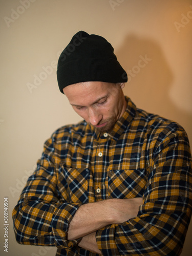 Young man, model of fashion, wearing a plaid shirt with a biege blind behind him. Guy in black hat with beard in casual style.