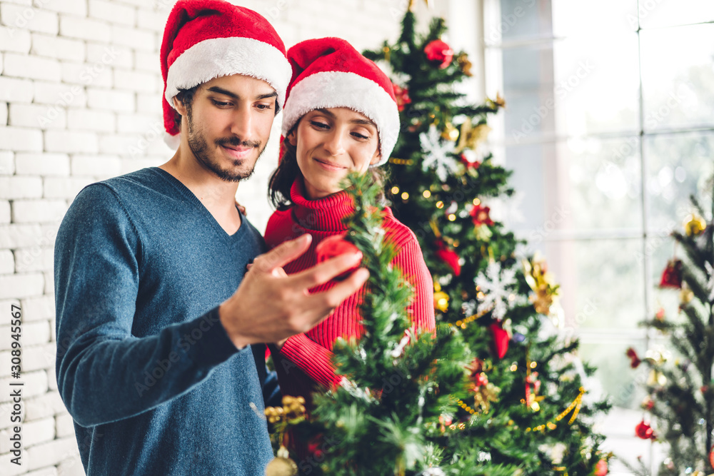 Romantic sweet couple in santa hats having fun decorating christmas tree and smiling while celebrating new year eve and enjoying spending time together in christmas time at home Romantic sweet couple in santa hats having fun decorating christmas tree and smiling while celebrating new year eve and enjoying spending time together in christmas time at home