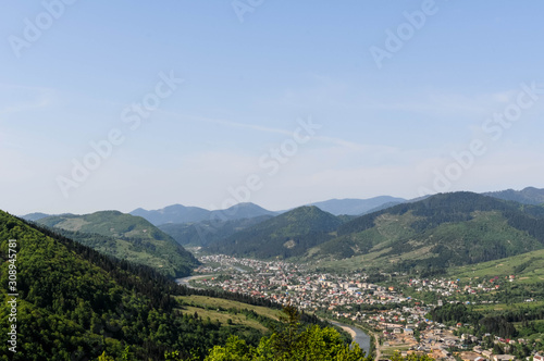 landscape of blue sky and town near big river in valley among mountains covered green forest © Viktoriia