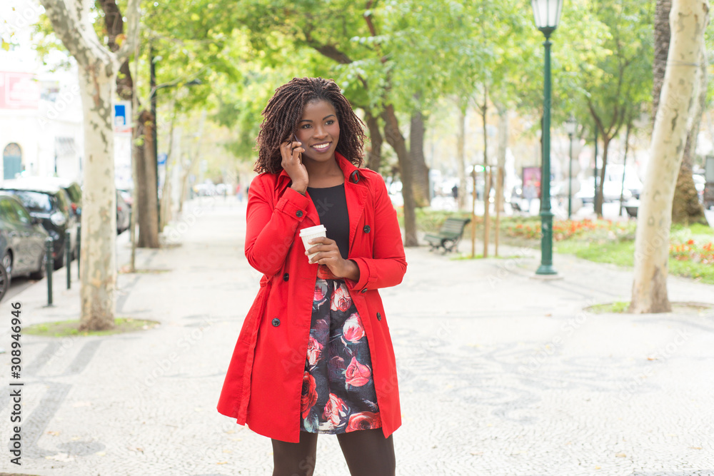 Content woman with coffee to go talking by smartphone. Beautiful smiling young African American woman holding paper cup and talking by cell phone in park. Connection concept