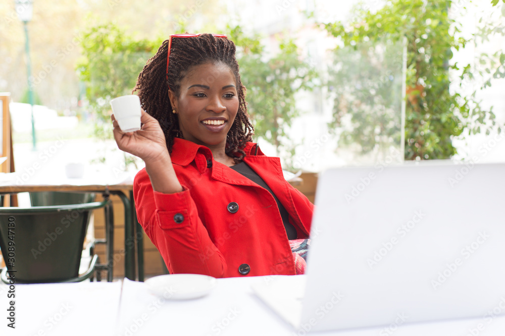 Content young woman using laptop in outdoor cafe. Beautiful smiling young African American woman holding cup of coffee and working with laptop in restaurant. Freelance concept Content young woman using laptop in outdoor cafe. Beautiful smiling young African American woman holding cup of coffee and working with laptop in restaurant. Freelance concept