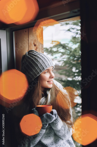 Beautiful woman holding and drinking a cup of coffee or cocoa in gloves sitting home by the window. Blurred winter snow tree background. Morning, coziness and people concept