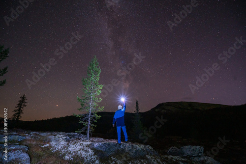 Milky way and starry sky over night scene outdoors in the forest and the mountains with man in blue clothes and flashlight in the foreground. Landscape, astronomy and wilderness concept.