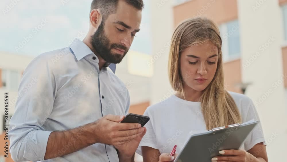 Calm pensive young office colleagues counting something with smartphone and writing in clipboard while standing outdoors Calm pensive young office colleagues counting something with smartphone and writing in clipboard while standing outdoors