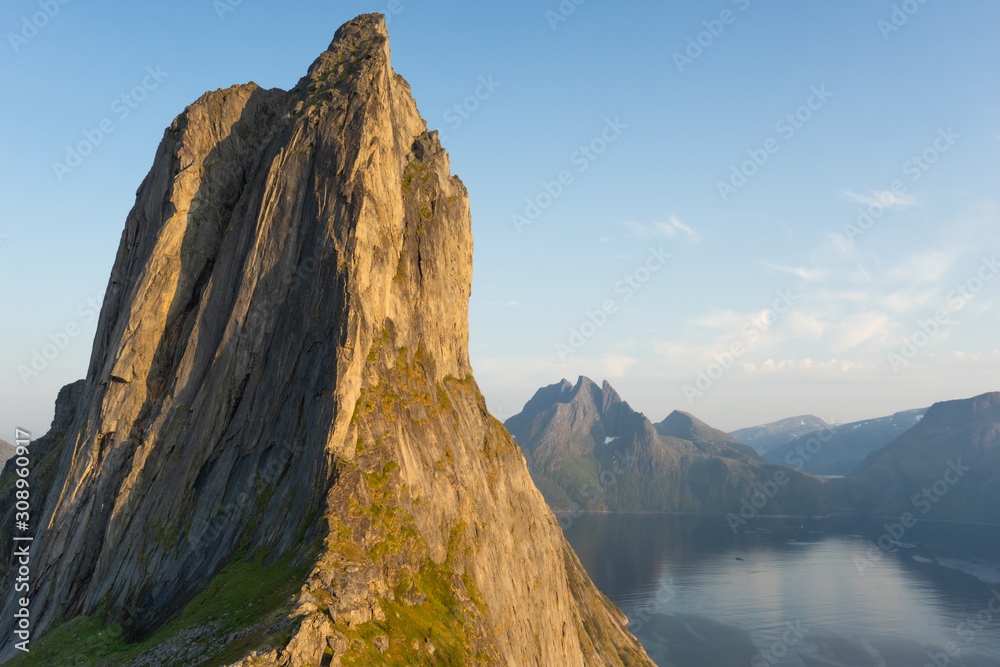Cliff wall bathed in evening sun. The mountain top Segla shaped as a sail. In the background the Norwegian fjord Mefjord and mountains. Cliff wall bathed in evening sun. The mountain top Segla shaped as a sail. In the background the Norwegian fjord Mefjord and mountains.