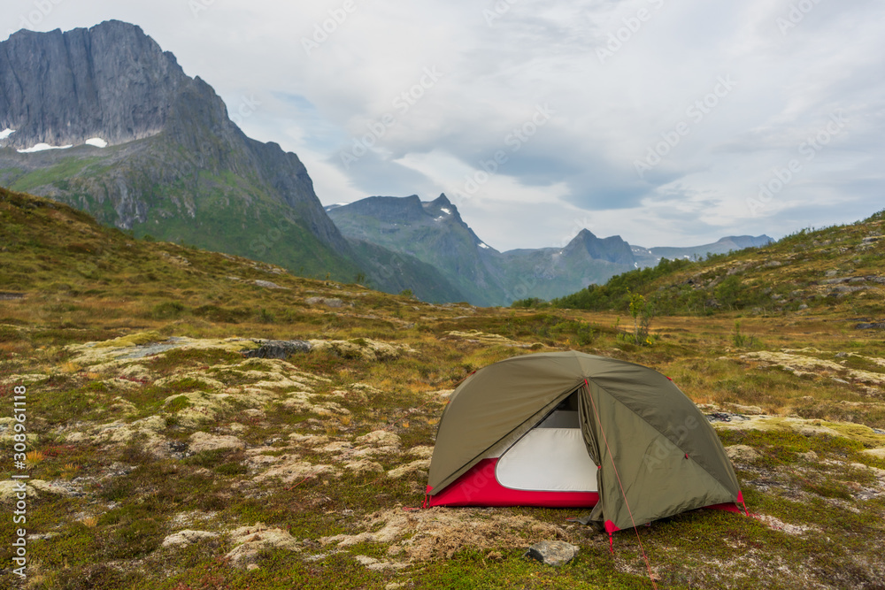 Open tent in green and red colors standing in wild landscape with mountain tops in the background. Autumn weather and clouds on the sky. Senja, Norway.