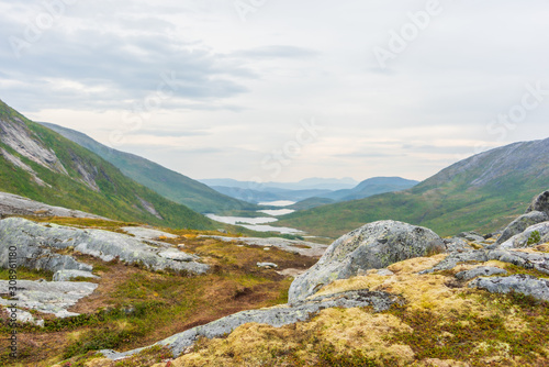 View from a high perspective over green mountain landscape with lakes in a valley. In foreground a rocky plateau with yellow colors. Senja, Norway.
