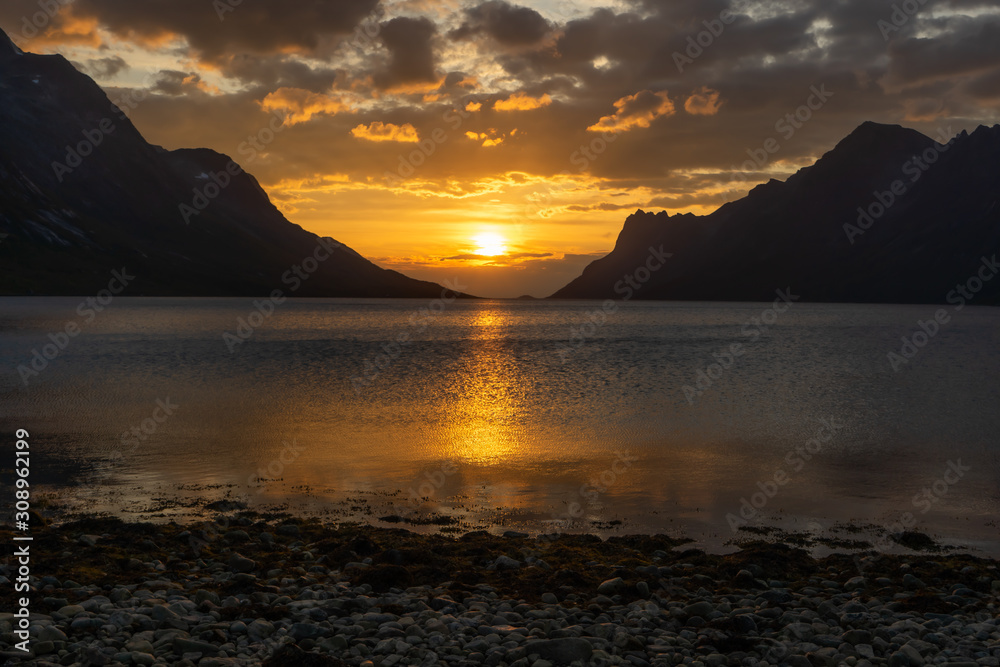 Fototapeta premium Golden sunset at Norwegian fjord with mountains. Sun shines on clouds above and reflects in the water. Dark mountain silhouttes. In foreground stone shore. Ersfjordbotn, Tromso, Norway.