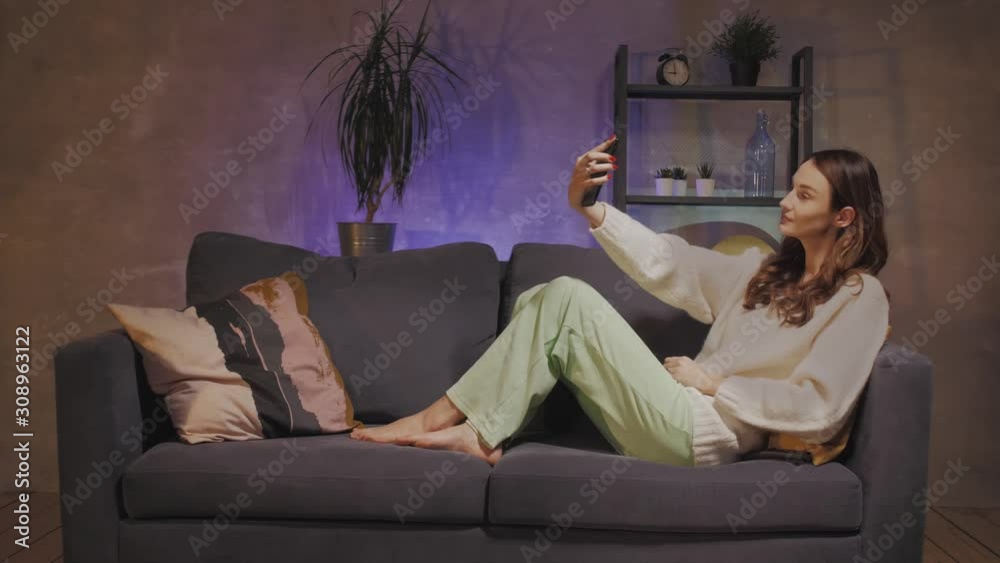 A young woman sitting on a sofa in a cozy room takes a selfie and smiles A young woman sitting on a sofa in a cozy room takes a selfie and smiles