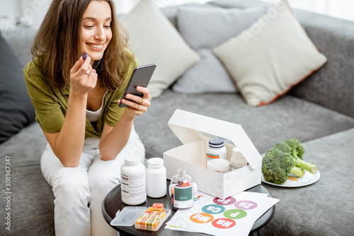 Woman taking nutritional supplements in the form of pills while sitting with smart phone at home. Concept of individual online selection of food supplements. Preventive medicine photo
