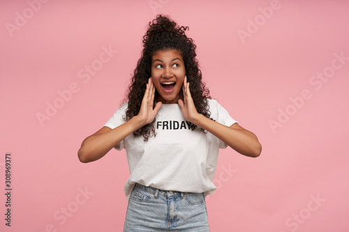 Cheerful attractive young brunette dark skinned female with long curly hair looking happily aside and touching her face with fingers, keeping her mouth wide opened while posing over pink background