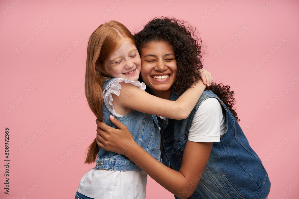 Studio portrait of happy cute girls enjoying gentle hugs while posing over pink background, smiling cheerfully and keeping eyes closed, wearing jeans vests and white shirts Studio portrait of happy cute girls enjoying gentle hugs while posing over pink background, smiling cheerfully and keeping eyes closed, wearing jeans vests and white shirts