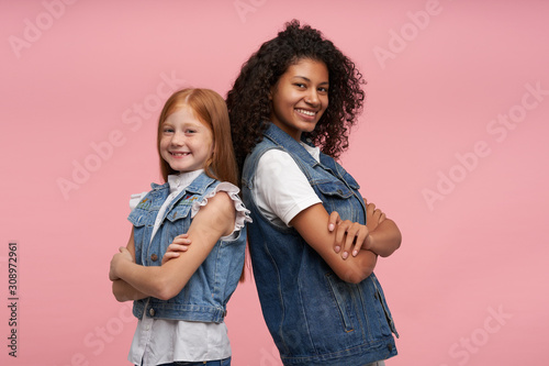 Studio shot of cheerful pretty young girls leaning on each other and smiling happily to camera with broad smiles, keeping hands folded while posing over pink background